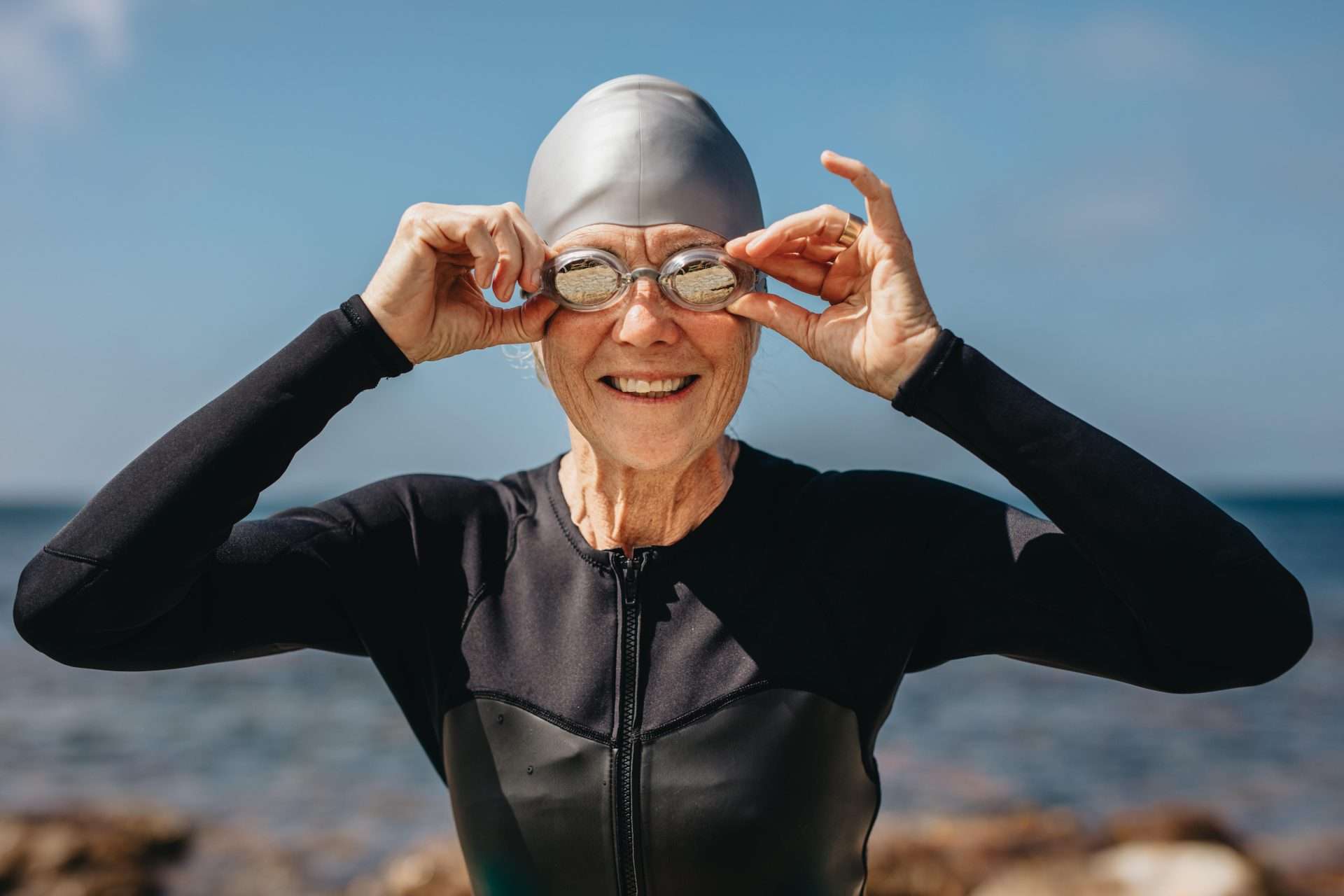 Portrait of a smiling senior female swimmer with sea in the background. Cheerful senior woman in swim wear standing on the beach holding her swimming
